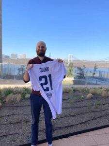 Dr Pete Quinn holding a white football jersey with his last name and #21 on it in blue