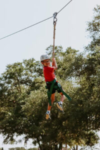 Young boy wearing a helmet, smiling as he holds onto a harness and wire on a zip line above the ground
