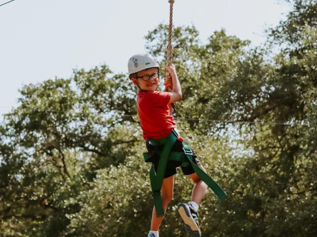 Young boy wearing a helmet, smiling as he holds onto a harness and wire on a zip line above the ground