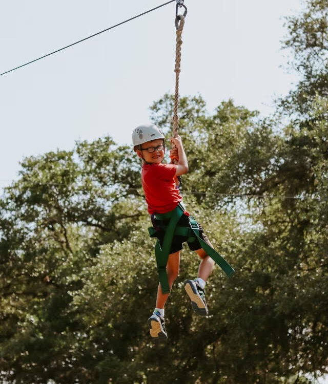 Young boy wearing a helmet, smiling as he holds onto a harness and wire on a zip line above the ground